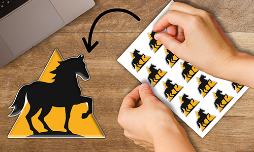 Hand peeling off horse silhouette sticker from sheet — yellow triangle design, one sticker placed near laptop.