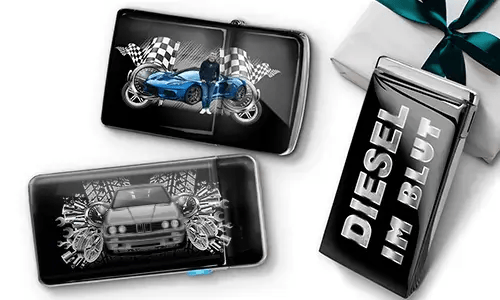 Two sleek black lighters featuring car themes, one with a blue car and checkered flags, another with a red car and gears. A third lighter reads 'DIESEL IM BLUT'. Gift-wrapped in the background.
