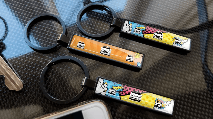 Porte-clés style bande dessinée avec plaque d’immatriculation et illustrations de voitures colorées, exposés sur une surface à texture carbone
